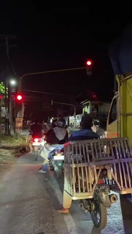 The video captures a nighttime street scene from the perspective of someone on a motorcycle, observing traffic at an intersection. The focal point is a traffic light that cycles from red to green. Several motorcycles are stopped at the intersection, facing forward. One motorcycle in the foreground has a wooden cage attached to its rear, containing what appear to be animals, possibly poultry. The rider of this motorcycle is wearing a blue shirt and light-colored pants, with their bare feet visible. Behind this motorcycle, other riders are also waiting. A large yellow truck is positioned to the right of the frame, its side visible. The street is paved, and there are streetlights and overhead wires present. The background shows buildings and trees, illuminated by streetlights and the red glow of the traffic signal. The overall atmosphere is that of a typical urban or semi-urban intersection at night.