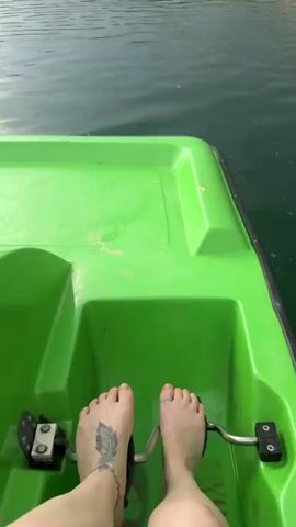 The video shows a first-person view from a green paddle boat on a body of water. The water has ripples and appears to be a dark green color. In the foreground, the edge of the green boat is visible, and a bare foot is resting on the boat floor. In the background, there is a landscape with a sandy cliff face covered in green trees. The sky is overcast with dramatic clouds. Further in the distance, there are buildings and construction cranes visible on the horizon. The boat appears to be moving across the water, as indicated by the shifting perspective of the background. A single person on a paddleboard is visible in the distance.