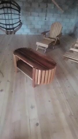 The video shows an indoor scene with wooden furniture. In the foreground, there is an oval-shaped wooden coffee table with a lower shelf. The table is made of various shades of wood, with slatted sides and a top composed of lengthwise planks. Behind the coffee table, two Adirondack chairs are visible. The chair on the left is fully in view, while the chair on the right is partially visible. The furniture is placed on a light-colored wooden floor. The background features a brick wall and what appears to be a wooden rack hanging on the wall. There are also some electrical cords on the floor. The lighting is somewhat dim.