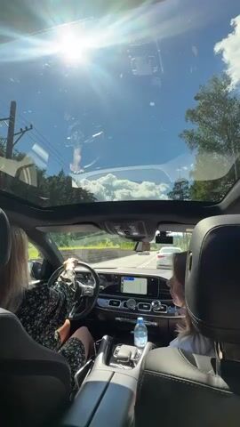 This is a first-person perspective video taken from inside a car, looking forward through the windshield and a sunroof. The sun is shining brightly, creating lens flares across the image. The sky is blue with scattered white clouds. We see the dashboard of the car, including a navigation screen. A woman is visible driving, with her hands on the steering wheel. Her back is to the camera, and she is wearing a dark, floral-patterned dress. Another person's head is visible in the passenger seat, also with their back to the camera. Outside the car, we see a road with other vehicles, trees, and some buildings. The car appears to be in motion.