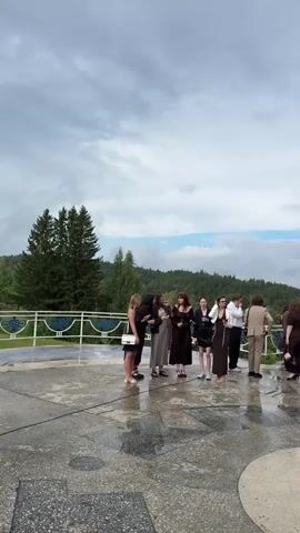 A group of people are gathered on a paved outdoor area, possibly a balcony or terrace, overlooking a body of water and a forested landscape. The sky is overcast with clouds. The pavement is wet, suggesting it has rained recently.

In the foreground, a group of approximately ten people, dressed in formal attire, are standing and interacting. Some are women in dresses, and some are men in suits. One woman on the left is wearing a dark dress and holding a light-colored clutch. Another woman in the center is wearing a long, dark brown dress and holding a dark purse. A man in a tan suit is standing slightly apart from the main group.

To the right, a wedding arch decorated with greenery and white fabric is visible. Two people in formal wear are standing near the arch. Further to the right, another group of people are gathered, including a man in a dark suit holding a camera.

In the background, trees line the edge of the terrace, and beyond that, a wide body of water is visible, with a distant shore and a hazy sky. Another group of people are standing near the railing, some observing the scene. A photographer in a black shirt and pants is standing in the middle ground, looking down at his camera.

As the video progresses, the camera pans to the right, showing a woman in a light-colored dress walking towards the group with flowers. Another person is seen holding a bouquet of white flowers wrapped in purple paper. The final shot shows the photographer again, now in the foreground, with the paved area and trees in the background. The overall atmosphere appears to be a wedding or special event, possibly taking place outdoors despite the cloudy weather.