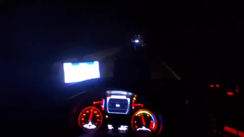 The video shows the dashboard perspective of a motorcycle driving on a dark road at night. The dashboard screen and speedometer are visible, along with the headlights of other vehicles in the distance. Music is playing in the background.