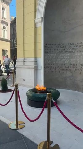 The video shows a view of an eternal flame memorial in front of a building with a large arched entryway. The flame is situated in a circular green base. The wall behind the flame has text inscribed on it, which appears to be a memorial inscription. Red velvet ropes on golden stanchions are set up in front of the memorial, creating a barrier. To the left of the memorial, a woman walks past, and further in the background, two men are visible near a car. On the building above the arched entryway, a banner with "Creative Europe" is displayed, along with the European Union flag. The environment appears to be an urban setting with buildings and a street visible in the background. The weather is sunny.