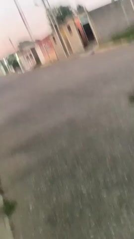 The video shows a street corner in a residential area, possibly at dusk. A grey car is parked on the right side of the street. Streetlights illuminate the area. The sky is a mix of pale orange and white. Power lines are strung across the sky. There is a tall cell tower. Houses are visible on either side of the street, and the camera is angled upwards. The camera seems to be hand held.