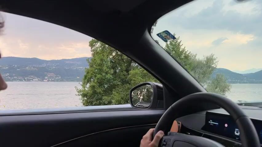 The video is shot from inside a car, looking out the passenger side window and through the windshield. A person's head and shoulder are visible on the left side of the frame, facing forward. The car is driving along a road next to a large body of water, likely a lake, with mountains and trees in the background. The water is calm, and there are a few boats visible. The sky is cloudy. In the middle of the video, two people wearing helmets are seen on the right side of the frame, standing near the road. The car has a digital dashboard visible in the lower right corner.