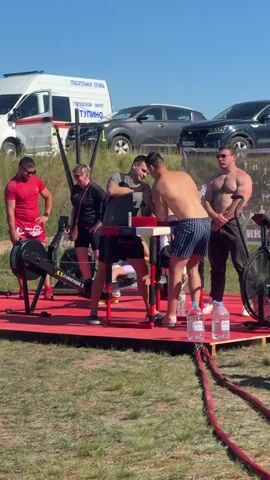 Two men are arm wrestling on a red platform outdoors. Several other people are standing around them, watching. There are exercise machines, water bottles, and vehicles in the background. The sky is blue and the sun is shining.