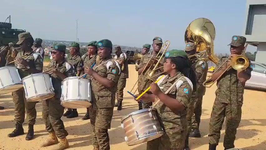 A military band, composed of approximately 15 individuals, is shown performing outdoors. The band members are dressed in camouflage uniforms and green berets. They are playing various instruments, including drums, trombones, and a tuba. The drums are white and appear to be bass drums and snare drums. The brass instruments are gold in color. Some members are using drumsticks, while others are playing their instruments. The individuals are arranged in rows, with the drummers in the front and the brass players behind them. The setting is outdoors, with a dirt ground and a clear sky. In the background, a military vehicle and buildings can be seen. The lighting suggests it is daytime.