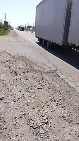 The video shows a roadside view of a multi-lane highway on a bright sunny day. The highway is cracked in places, showing signs of disrepair. Several large semi-trucks are driving down the road, in the same direction as the camera. The shoulder of the road is gravelly. There are electric power lines visible on the far side of the road.