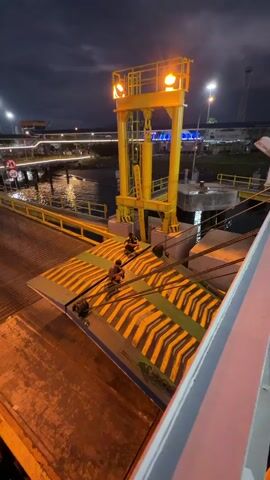 The video shows a night scene at a dock or ferry terminal. Two individuals are visible on a yellow and black striped ramp or walkway that extends from a larger structure, likely a ferry or loading platform, towards the shore. The ramp is illuminated by artificial lights, casting a warm glow. In the background, a tall yellow gantry or lifting structure with lights is prominent. The water of the harbor is visible to the left, reflecting the lights of the surrounding area, which includes buildings and other dockside infrastructure. The sky is dark and appears to be cloudy. One person is kneeling on the ramp, while the other stands nearby, looking towards the shore. The overall impression is of a port operation in progress at night.