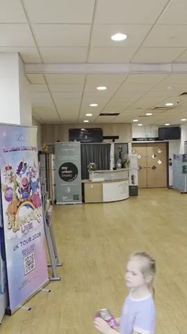 The video shows the interior of a Leisure World facility. The camera pans from left to right, revealing the reception desk, a door leading to a Charter Hall, a shop with leisure products, and a seating area with a sign thanking visitors. Several people are walking in the background. A small child with a backpack walks past the camera. The environment is brightly lit with fluorescent ceiling lights. The floor is made of light-colored wood. The overall atmosphere seems to be calm.