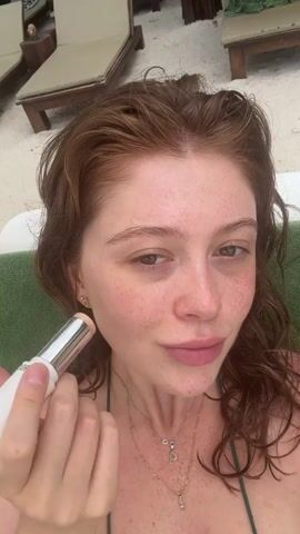 The media file features a close-up shot of a young woman with red hair, freckles, and light-colored eyes. She is wearing a green bikini top and a delicate necklace with a pendant. The woman is outdoors, likely in a tropical setting, as indicated by the background which shows lounge chairs on a sandy beach and some greenery. The lighting suggests it is daytime, possibly morning or afternoon, with no harsh shadows.

The woman is holding a white cylindrical object with a metallic top, identified as a Dior product, and is applying it to her cheekbone with a circular motion. Her expression is relaxed and natural, suggesting she is comfortable in her surroundings and perhaps enjoying a moment of self-care. The overall mood is serene and casual, fitting for a vacation or a relaxed outdoor setting like Ao Cho, Thailand. The visible text on the object reads "CHRISTIAN DIOR."