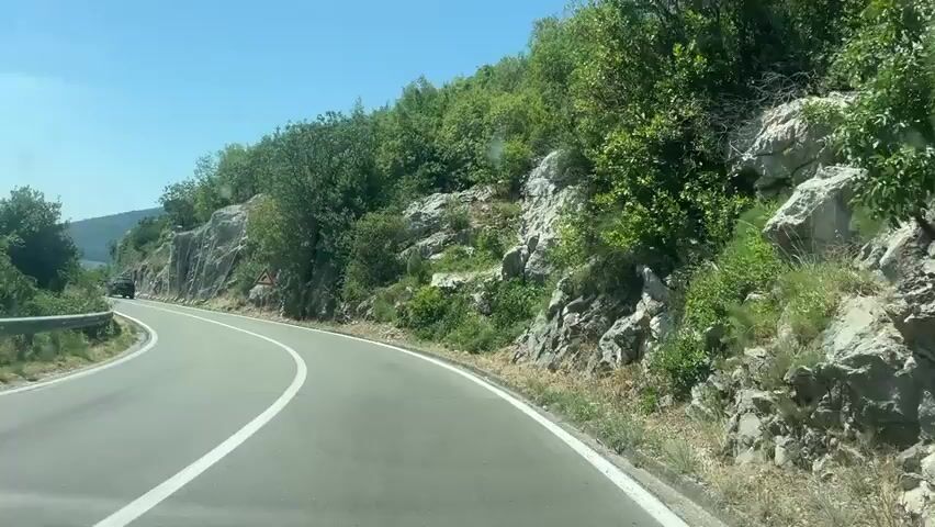The video shows a first-person view of driving on a winding road. The road is paved with white lane markings. To the left, there is a steep, rocky slope covered in vegetation. A guardrail lines the edge of the road on the left. In the distance, mountains are visible. On the right side of the road, there is a rocky cliff face that appears to be reinforced with a wire mesh. There are several trees and bushes growing on and around the rocky slopes. The sky is clear and blue. A few cars are visible on the road ahead. A triangular warning sign with a curve symbol is visible on the right side of the road.