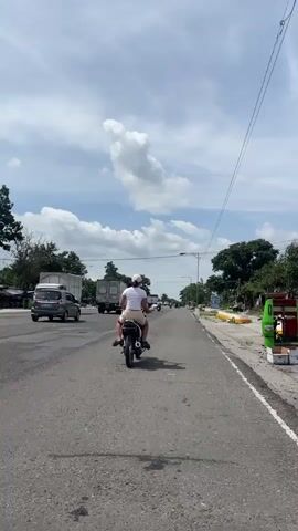 The media file captures a sunny day in Tupi, Philippines, with a view of a two-lane asphalt road. The sky is bright with scattered white clouds, suggesting it's daytime. Lush green trees line the sides of the road, and some small structures are visible in the distance, possibly shops or houses.

The primary subject is a person riding a motorcycle from behind, wearing a white t-shirt and shorts. Another person is seated behind the rider. The motorcycle is moving away from the camera, down the road.

Several vehicles are present on the road, including a white truck, a gray sedan, and another truck further down the road, indicating moderate traffic. The road appears to be in a semi-urban or rural setting, given the presence of trees, open space, and the style of buildings.

The overall mood appears casual and ordinary, depicting a common scene of transportation on a warm, clear day. There is no visible text within the frame.