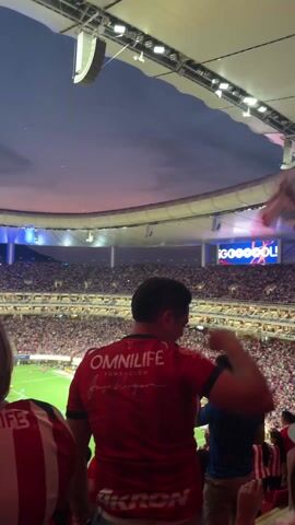 The video shows a view from the stands inside a large stadium filled with spectators. A soccer game is in progress below, though most of the view is of the packed stands and the stadium's lighting. In the foreground, a man wearing a red shirt with "OMNILIFE" and "AKRON" logos is facing the field. To his right, another man in a blue shirt stands. The stadium's curved roof is visible above, with a row of lights running along its edge. Large video screens display vibrant patterns and text. The overall atmosphere is energetic, with people moving and the stadium lit with alternating red and blue lights.
