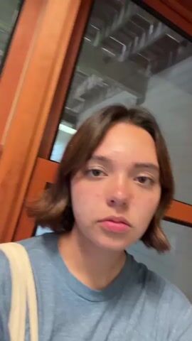 This media file features a young person with shoulder-length brown hair and fair skin, adorned with freckles. They are wearing a blue t-shirt with a white graphic design on the front. The person is holding a bundle of light-colored fabric in their arms and carrying a light-colored tote bag over their left shoulder. They appear to be indoors, near a window with wooden framing. The lighting suggests it is daytime, possibly late afternoon or early evening, given the soft, diffused light. The background visible through the window is slightly blurred, but hints at an outdoor environment with greenery. The person's facial expression is neutral to slightly discontent. The overall scene is casual and personal, possibly a moment captured spontaneously. No specific text is clearly legible.