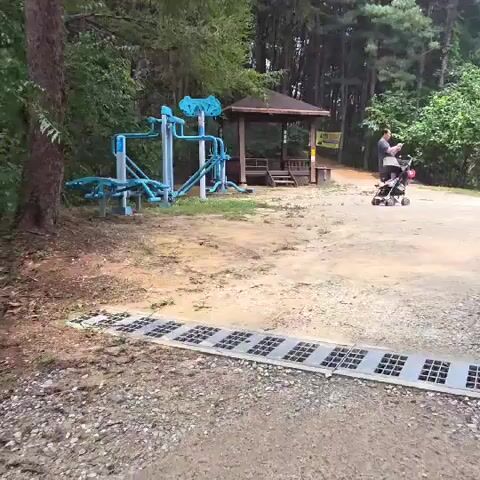 The video begins by showing an outdoor fitness equipment area, a wooden gazebo, and a dirt path. A person is pushing a stroller with a child inside along the path. The camera then pans to show a fork in the path with various signs and a barrier. The video continues to pan, showing more of the path and the surrounding wooded area. The scene appears to be a park or recreational area.
