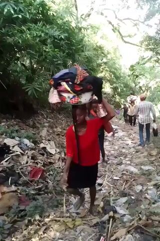 The video shows a group of people walking along a dirt path in a wooded area. The path is littered with debris and fallen leaves. The people in the video are carrying items on their heads, including bundles of clothing. The video is shot from a low angle, and the camera is shaky.