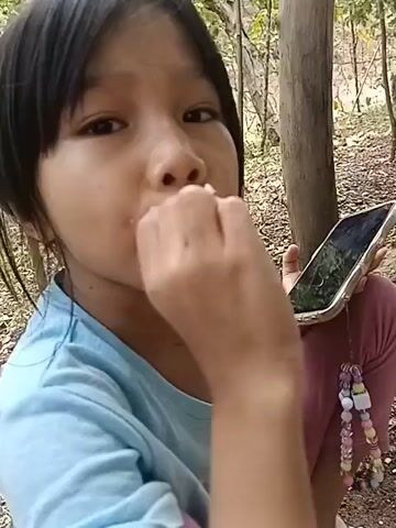 A young girl with dark hair is sitting outdoors, wearing a blue t-shirt and pink leggings. She is holding a smartphone in her right hand and appears to be eating from a pink plate in front of her. The plate contains rice, a piece of fish, and some green vegetables. The background shows trees and dry ground. The girl is looking towards the camera.