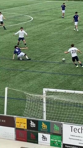 The video shows a soccer game. Two players, one wearing number 23 in a white jersey and the other wearing number 3 in a blue jersey, are close to each other near the lower left corner of the frame. They are both focused on the soccer ball. The player in the blue jersey is trying to take the ball from the player in the white jersey. Other players in similar jerseys are scattered around the green field. A white soccer goal net and a billboard advertising "Technotex" are visible in the foreground.