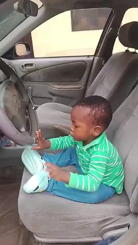 Inside a car, a small child sits on the driver's seat, dressed in a green and white striped shirt and blue jeans. The child, who is light-skinned with short black hair, is turned slightly to the left, looking at and touching the steering wheel. The car's interior is grey, with visible wear and tear on the seat fabric. Outside the car window, a building is partially visible.