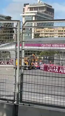 The video shows a Formula 1 race through a chain link fence. A yellow construction vehicle is working on the track behind a Qatar Airways banner. Formula 1 cars in various colors are visible, including pink and red. The sound of the engines is prominent. High-rise buildings are in the background against a partly cloudy sky.