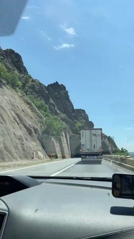 A forward-facing dashboard camera view shows a gray semi-trailer truck entering a tunnel. The tunnel is arched and constructed with large, rough-hewn stones, suggesting an older, well-built structure. To the left, a steep, rocky mountain face rises, covered in patches of green vegetation and some trees. The sky above is clear and bright blue, with a few wispy white clouds. The road is asphalt, marked with white lines, and appears to be a single lane in the direction of travel, with what might be an oncoming lane to the left. The truck's trailer is white with some red text or logos on its back doors. The dashboard is visible in the foreground, indicating the video is shot from inside a vehicle. There is a small screen or device mounted on the right side of the dashboard, and a digital display showing "30 C" can be seen on the left. The overall impression is a sunny day on a mountain road with a prominent tunnel entrance.