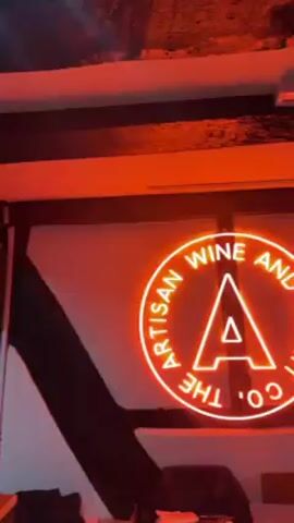 The video shows the interior of a small liquor store. The store has exposed beams on the ceiling, painted black. The walls are mostly white, with some black diagonal wooden supports.  A bright red neon sign on the wall reads "The Artisan Wine &".  Shelves of liquor bottles line the back wall, in front of the register. There are boxes of goods to the left side of the frame.
