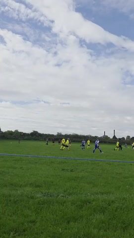 The video shows a soccer game on a grassy field under a cloudy sky. Several young players in yellow and blue uniforms are scattered across the field. In the foreground, a person with dark hair is partially visible on the right. The game appears to be in progress, with players moving and interacting.