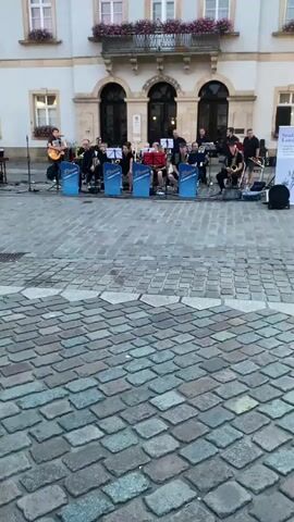 A jazz band performs on a cobblestone plaza in front of a building with arched windows and a balcony. The band consists of about ten musicians playing various instruments, including saxophones, trombones, and a guitar. They are seated behind blue screens with "Brass Connection" written on them. Several band members are looking at music stands. The building appears to be a public institution, possibly a town hall, given its architectural style. The scene is outdoors, and the lighting suggests it is either early morning or late afternoon.