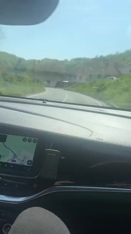 A car is driving on a rural road. The driver’s view is visible through the windshield. The dashboard, navigation screen, and steering wheel are visible. Other cars and houses are seen outside the car. The weather appears clear and sunny.