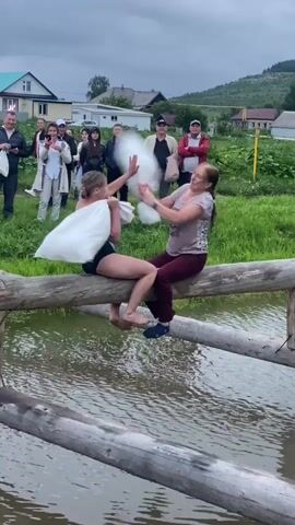 Two women are sitting on a log over water, facing each other. They are each holding a pillow and appear to be engaged in a pillow fight. A group of spectators is watching them from the grassy bank behind them. The environment appears to be rural, with houses and trees visible in the background under an overcast sky. The woman on the left is wearing a black swimsuit, and the woman on the right is wearing a patterned shirt and maroon pants.