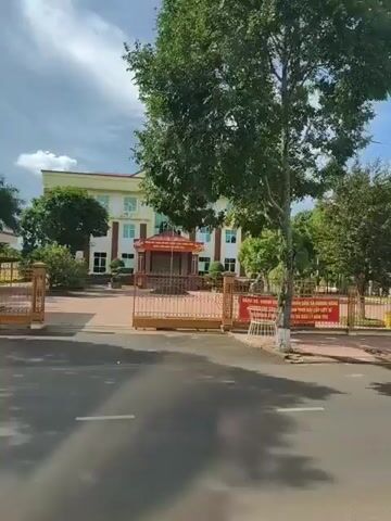 The video shows a street scene in front of a building. The building is light yellow with a green roof and appears to be a public building, possibly a government office or school. There are trees and greenery around the building. A gate is visible in front of the building, with banners displayed on it. A white truck with a dark blue cargo area drives past. Two motorcycles, one with two riders and one with a single rider, are seen driving on the road. The sky is cloudy. The footage appears to be taken from a moving vehicle.