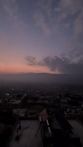 The video shows a cityscape at dusk. The sky is a mix of purple, blue, and peach, with clouds scattered throughout. Below, the city is dimly lit with scattered lights.  Buildings of varying sizes are visible, with flat rooftops dominating the foreground. One rooftop has what appear to be solar panels. Some hills are visible on the horizon. The scene is static with no visible movement or interaction.