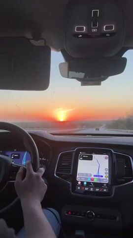 The video captures a serene sunset experienced from the inside of a car, likely a Volvo given the interior design. A person's hands are visible on the steering wheel, indicating they are driving.

The prominent feature is the vibrant orange and red sunset visible through the front windshield, with the sun low on the horizon directly ahead. A highway stretches into the distance, with other vehicles, including several trucks, also visible. The sky shows some faint contrails.

Inside the car, the dashboard and a large vertical infotainment screen are clearly visible. The screen displays navigation information, showing "139 km" remaining and a route on what appears to be highway "A5" towards a location indicated as "Hazinedar." The time displayed on the screen is "20:00," and temperature readings are "22°" and "24°". Various app icons are also present on the screen. The car's overhead console with passenger airbag indicators and lights is visible above the rearview mirror. A sun visor is lowered on the left side.

The overall scene conveys a peaceful long-distance drive during twilight in Babaeski, Turkey.