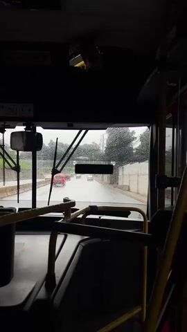 The video shows a view from inside a bus, looking out the windshield on a rainy day. Water droplets are visible on the windshield. The wipers are in the down position. The road ahead is wet, and several cars are driving in the same direction. Trees and foliage are visible on the sides of the road. The interior of the bus is dark, with yellow grab bars and black seats visible. The overall scene depicts travel in inclement weather.