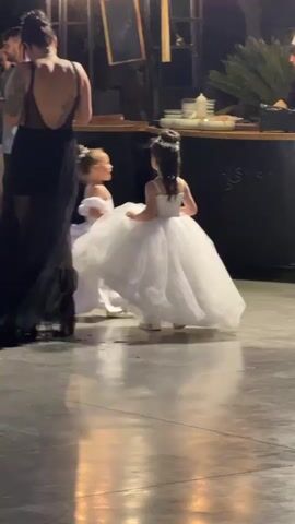 A group of people are gathered outdoors at night. In the foreground, two young girls in white dresses are dancing. A woman in a black dress is standing behind them. In the background, other people are socializing, and there are lights strung across the scene. The ground appears to be a polished concrete floor.