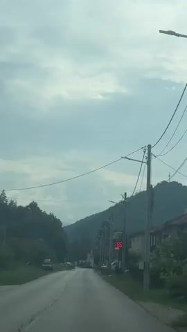 The video shows a view from a moving vehicle on a paved road. The road is flanked by trees and houses. Overhead power lines are visible, supported by utility poles. A traffic light is illuminated red. In the distance, a hill or mountain covered in trees rises against a cloudy sky. Several cars are parked or driving on the road. The overall impression is a rural or suburban environment on an overcast day.