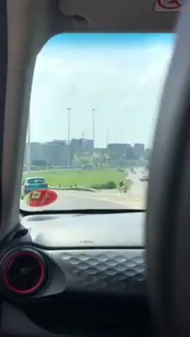 The video, shot from inside a moving car, shows a view through the windshield. We see a paved road with green medians on either side.  Buildings are visible in the distance under a bright, blue sky.  The car's dashboard is in the foreground, and an air vent with a red ring is visible. The car is driving on a sunny day.