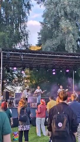 An outdoor concert is underway. A band is performing on a stage, with several members playing instruments and singing. A crowd of people is gathered in front of the stage, watching the performance. The scene is set in a grassy area with trees in the background. The lighting suggests it is either late afternoon or early evening.