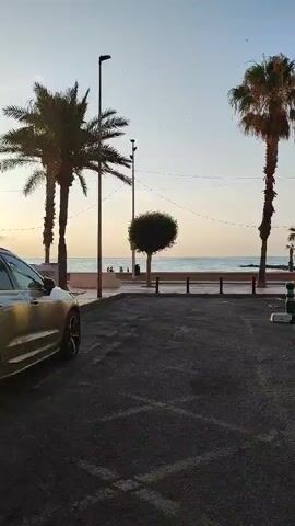 The video shows a parking lot with a car in the foreground. Behind the car is a beach scene with palm trees and the ocean. The sun is setting, casting a warm glow on the scene. There are a few people walking on the beach.