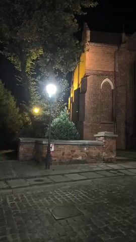 The video shows an exterior view of a brick building at night. A street lamp illuminates a section of the cobblestone ground and a low brick wall. Trees are visible to the left of the frame. The building appears to be a church or similar structure, with arched window shapes visible in the brickwork. The lighting suggests it is nighttime or a very dark environment. There are no people visible in the video.