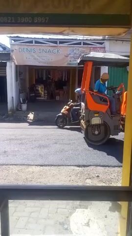 Here is a concise description of the video:

The video shows an orange and gray Hamm HD 30 asphalt roller moving along a newly paved road. The roller is operated by a man wearing a blue shirt and hat. A motorcyclist is visible on the left side of the road, passing the roller. In the background, there are storefronts including one with a sign that says "DENIS SNACK" and another advertising NutriSari. The video appears to be shot from inside a building or vehicle, looking out through a window. The camera is mostly stationary, with a slight shaking. The weather appears to be sunny.