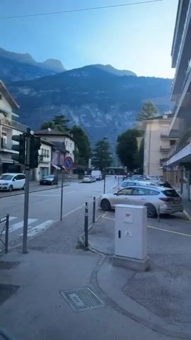 The video shows a street scene with mountains in the background and buildings lining the right side of the road. Cars are parked along the curb. A traffic light is visible on the left. The sun is shining, casting long shadows. The video pans slightly to the right, revealing more of the buildings and parked cars. A white van is parked on the left side of the street. A red and white no parking sign is posted on the right side of the street.