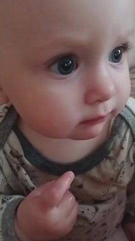 A close-up video of a baby's face. The baby has wide, blue eyes and fair skin. The baby is wearing a light-colored onesie with a grey collar. The baby's left hand is visible, with its index finger extended. The background is blurred and appears to be an indoor setting with a patterned fabric. The video is 4 seconds long and shows the baby looking to the right, then turning its head downwards.