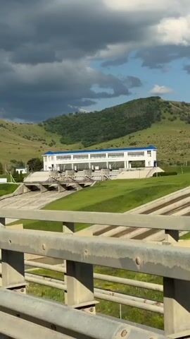 The video shows a concrete dam with a building on top, situated in a valley with green hills. The dam has multiple sluice gates, and water is flowing through them, creating a whitewater effect. The building is white with a blue roof and appears to be an administrative or control structure for the dam. The sky is partly cloudy, with dark clouds in the distance. In the foreground, there is a blurry metal railing, suggesting the video was filmed from a bridge or an elevated viewpoint. The overall scene is of a functional water management facility in a natural, hilly landscape.