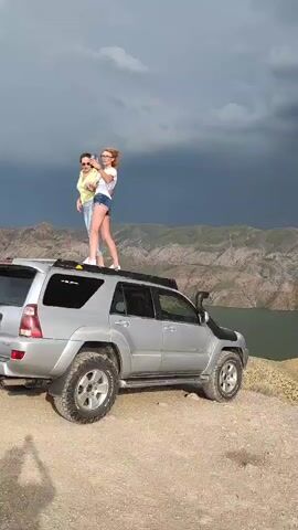 The video shows a silver SUV parked on a dirt surface with a body of water and mountains in the background. Two young women are standing on the roof of the SUV, taking a selfie with a smartphone. The sky is dark and overcast, suggesting an approaching storm. One woman is wearing a yellow top and blue jeans, while the other is wearing a white t-shirt and denim shorts. A man is seen walking in the background. The SUV has off-road tires and a snorkel attached to the front.