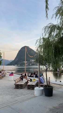 The video shows an outdoor seating area with people relaxing near a body of water, possibly a lake, with a mountain in the background. String lights are hung between poles, illuminating the area as dusk settles. There are several groups of people sitting on benches made from wooden pallets. Some individuals are talking to each other, while others appear to be looking at their phones. There are trees and potted plants scattered throughout the area, adding to the natural ambiance. The ground is covered in gravel and sand. The sky is a pale blue with scattered clouds, and the water reflects the lights from the shore and distant town. The overall atmosphere seems relaxed and social.