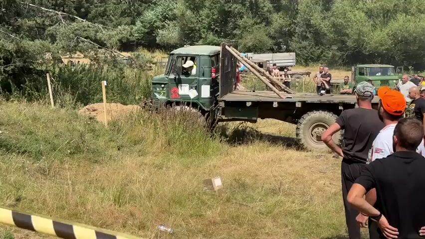 A group of people watch as a green off-road truck attempts to navigate a muddy incline. The truck is positioned in a grassy area with trees and other vehicles in the background. Some people are standing close to the truck, while others are observing from a distance. The truck appears to be stuck, with its front wheels dug into the mud. The people around the truck are not actively assisting it, but rather observing its progress. The overall atmosphere seems to be that of an outdoor event or competition.