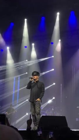 A man dressed in black stands on a stage illuminated by bright spotlights and blue lights, singing into a microphone. He is wearing a black button-down shirt, patterned black pants, and a hat. A guitarist is visible on the left side of the stage and a drum set can be seen behind the main figure. The audience holds up their phones to record the performance. The atmosphere suggests a concert or live music event.