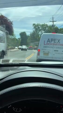 The video shows a view from inside a car, looking out through the windshield. The car is stopped in traffic on a two-lane road with a double yellow line. In front of the car is a white van with the words "APEX LOCKSMITHS EMERGENCY SERVICE" printed on its side. Further ahead, there is a police car with its lights flashing. Other cars can be seen in front of the police car. The environment appears to be a residential area with trees lining the road. The sky is partly cloudy.